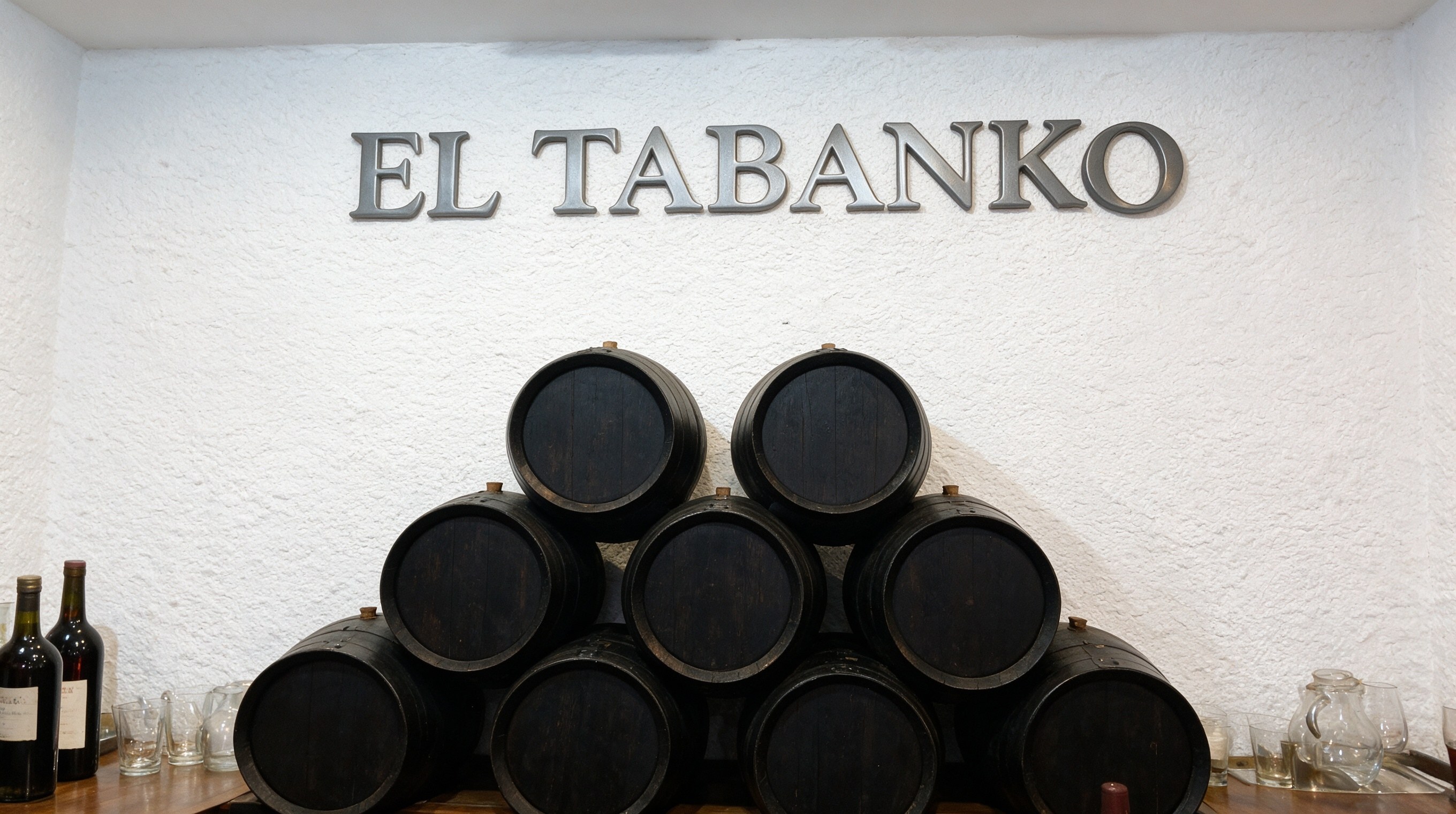 Pared blanca con rótulo en relieve EL TABANKO y botas de jerez apiladas en un tabanco